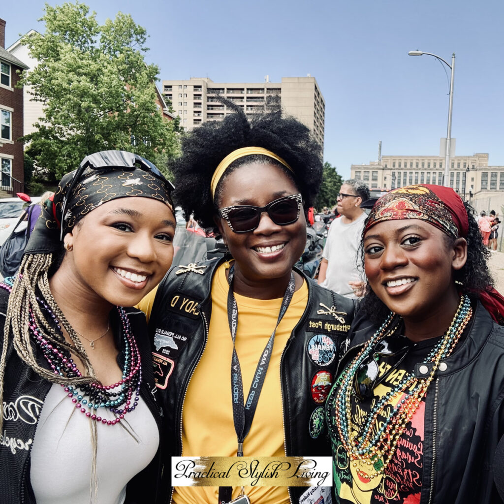 Motorcycle Riding Boots A group of three smiling women pose closely together for a bright, outdoor portrait during a daytime event. All three wear decorative headwraps and various layered necklaces, including colorful beads.