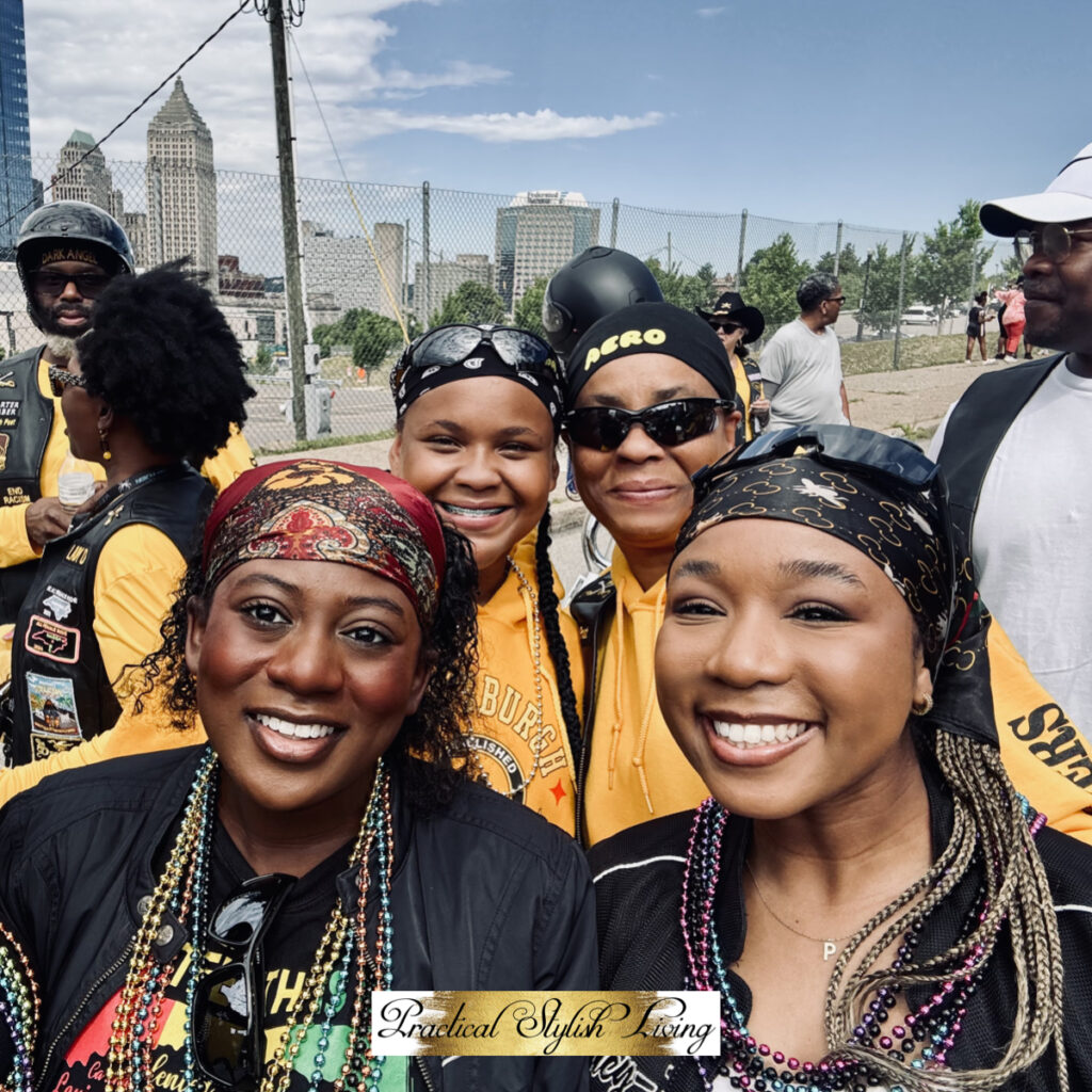 four smiling Black women at an outdoor gathering, possibly a festival or rally, with a city skyline behind a chain-link fence in the background. Most are wearing yellow clothing and decorative accessories like headwraps and multi-colored beads. A few men are also visible in the crowd, including one in a motorcycle helmet and leather vest.